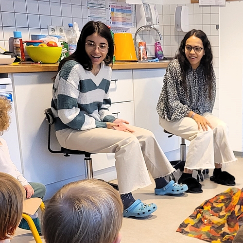 Zwei junge Frauen sitzen mit Kindern in einer Kita-Küche. Sie lachen und gestalten gemeinsam eine Aktivität mit bunten Tüchern auf dem Boden.