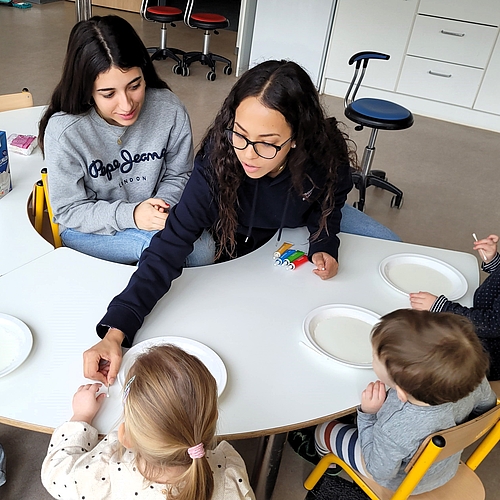 Zwei junge Frauen sitzen mit einer kleinen Kindergruppe an einem Tisch. Eine von ihnen zeigt einem Kind etwas mit einem Wattestäbchen auf einem Pappteller.